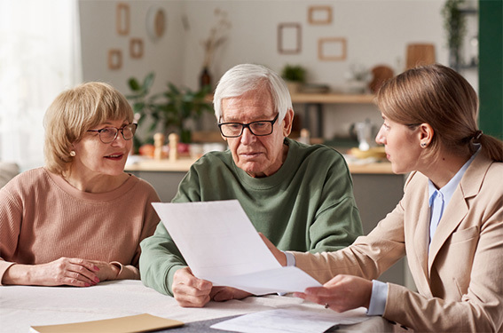 Older couple seated at a table with a professional advisor in a warm, home like setting. The advisor gestures towards documents while explaining information. The couple look attentive and engaged, reviewing the papers together. The scene conveys reassurance, trust and support while discussing important personal or financial matters.