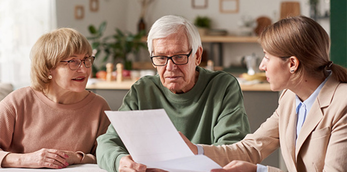 Older couple seated at a table with a professional advisor in a warm, home like setting. The advisor gestures towards documents while explaining information. The couple look attentive and engaged, reviewing the papers together. The scene conveys reassurance, trust and support while discussing important personal or financial matters.