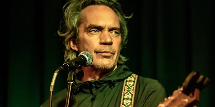 A musician performs on stage under warm lighting, playing an acoustic guitar and singing into a microphone. They wear a dark jacket and stand against a backdrop of deep green curtains. The scene has an intimate, atmospheric feel typical of a small live music venue.