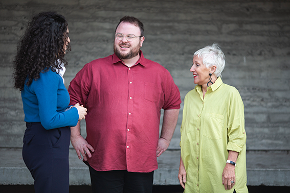 Three adults stand talking in front of a grey concrete wall. One person wears a blue top and dark trousers, another wears a red button-up shirt, and the third wears a light green shirt. The group faces each other in conversation in an outdoor setting.