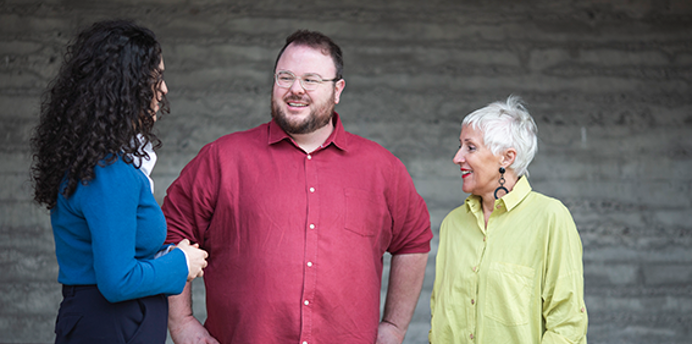 Three adults stand talking in front of a grey concrete wall. One person wears a blue top and dark trousers, another wears a red button-up shirt, and the third wears a light green shirt. The group faces each other in conversation in an outdoor setting.