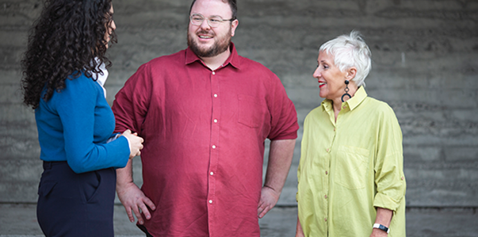 Three adults stand talking in front of a grey concrete wall. One person wears a blue top and dark trousers, another wears a red button-up shirt, and the third wears a light green shirt. The group faces each other in conversation in an outdoor setting.
