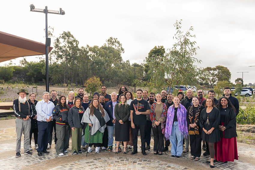 A group of approximately 30 people standing together outdoors in a landscaped gathering space, posing for a group photograph. The group is arranged in several rows on a paved circular area, with native trees, shrubs and open bushland visible in the background. A modern building structure is visible on the left, and a tall light pole stands behind the group. Participants are wearing a mix of casual, professional and culturally significant clothing. The photo was taken during daylight under an overcast sky.