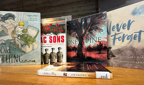 A display of books about ANZAC Day and Australian wartime history standing on a wooden surface. Visible titles include This Old Thing, AC Sons on the Western Front, One Pine, Never Forget, and Soldier Boy by Anthony Hill.