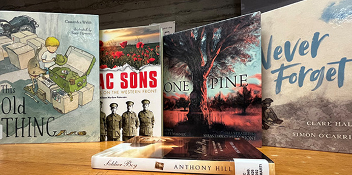 A display of books about ANZAC Day and Australian wartime history standing on a wooden surface. Visible titles include This Old Thing, AC Sons on the Western Front, One Pine, Never Forget, and Soldier Boy by Anthony Hill.