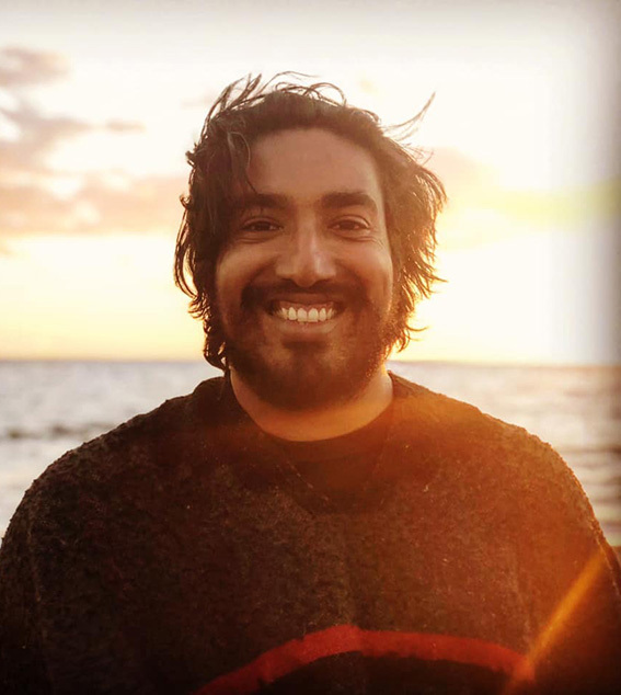 Portrait photograph of a person standing outdoors near the ocean at sunset. The person is facing the camera and wearing a dark top. The background shows water and a warm, glowing sky with sunlight near the horizon.