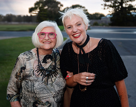 Two people standing close together outdoors, smiling at the camera. One wears patterned clothing with bright glasses, the other wears a dark textured outfit with light accessories. Both appear well dressed and confident. They stand on grass near a road with trees and open space behind them, photographed in soft natural light that gives the image a warm, welcoming feel.