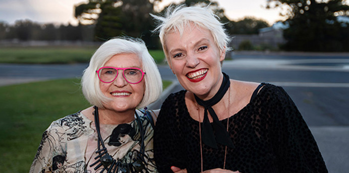 Two people standing close together outdoors, smiling at the camera. One wears patterned clothing with bright glasses, the other wears a dark textured outfit with light accessories. Both appear well dressed and confident. They stand on grass near a road with trees and open space behind them, photographed in soft natural light that gives the image a warm, welcoming feel.