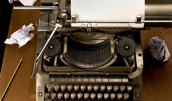 Top‑down view of a vintage typewriter on a wooden desk, with a blank sheet of paper loaded, a pencil beside it, and several crumpled pieces of paper scattered around, suggesting the process of writing and rewriting.
