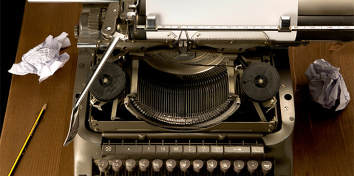 Top‑down view of a vintage typewriter on a wooden desk, with a blank sheet of paper loaded, a pencil beside it, and several crumpled pieces of paper scattered around, suggesting the process of writing and rewriting.
