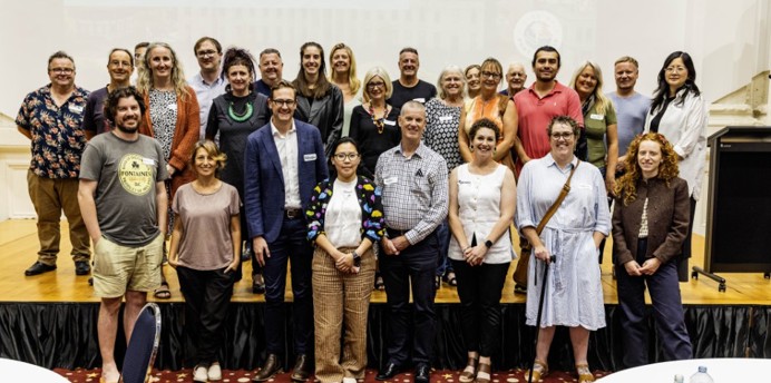 A large group of people stands together on a stage in a function room, posing for a group photo. They are dressed in a mix of business and smart‑casual clothing. The background shows a projection screen with the words “Welcome and Acknowledgment of Country”. The room features patterned carpet, round tables and a podium, creating a formal community or organisational event setting.